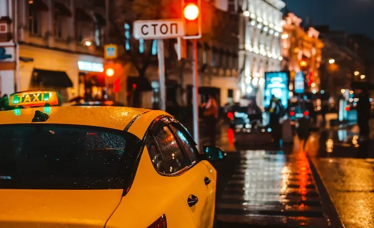 A yellow taxi stopped on a wet city street at night, shop lights reflected in the tarmac